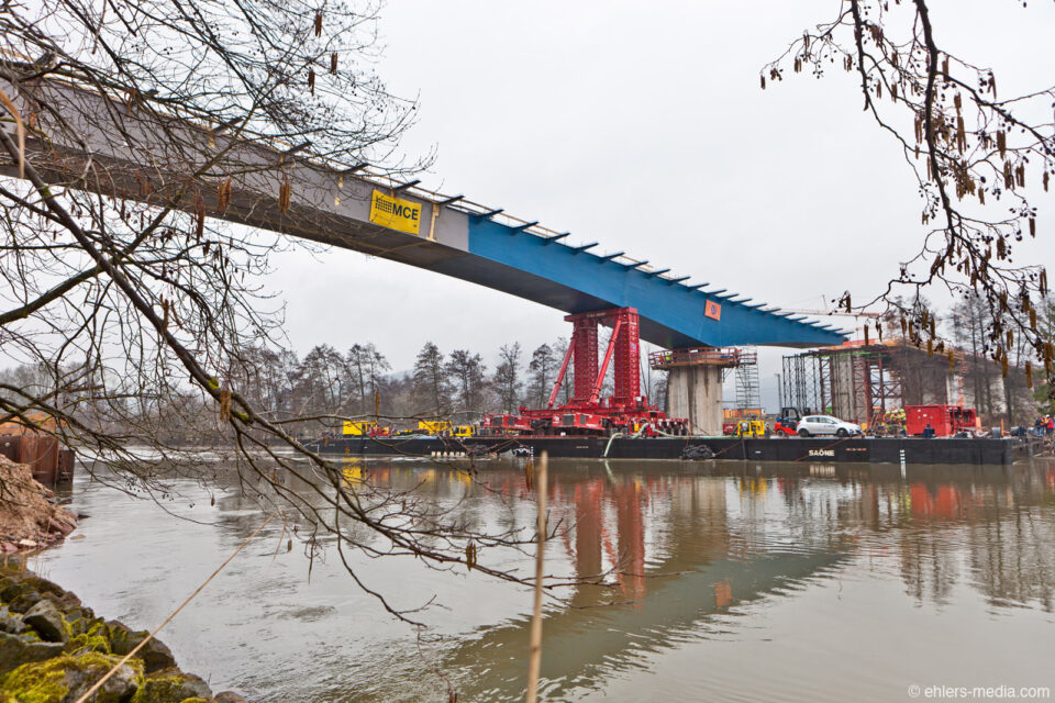 Brückenbaustelle über einem Gewässer mit teilweise montierten blauen und grauen Brückensegmenten; Kräne, Baumaschinen und Arbeiter sind sichtbar, eingerahmt von Bäumen und Ästen in natürlicher Umgebung.