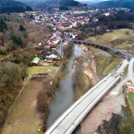 Luftaufnahme einer Kleinstadt mit Flusslauf und mehreren Brücken, darunter eine im Bau; umgeben von hügeliger Landschaft, Wäldern, Wohnhäusern, Straßen und offenen Feldern.