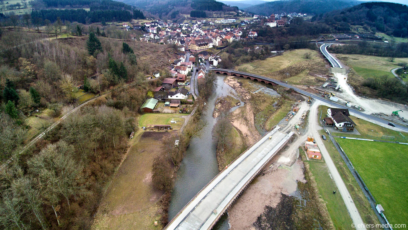 Luftaufnahme einer Kleinstadt mit Flusslauf und mehreren Brücken, darunter eine im Bau; umgeben von hügeliger Landschaft, Wäldern, Wohnhäusern, Straßen und offenen Feldern.
