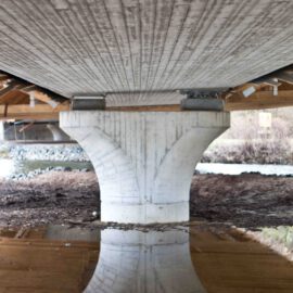 Unterseite einer Brücke im Bau mit zentralem Betonpfeiler, dessen Spiegelung im Wasser darunter sichtbar ist.