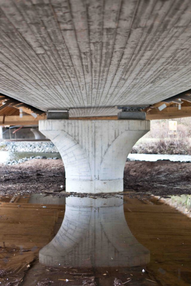 Unterseite einer Brücke im Bau mit zentralem Betonpfeiler, dessen Spiegelung im Wasser darunter sichtbar ist.