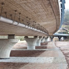 Unterseite einer Brücke mit mehreren Betonpfeilern, an der Metallrohre und Kabel entlanglaufen; darunter teils befestigter, teils unbefestigter Boden, im Hintergrund Bäume und Vegetation.