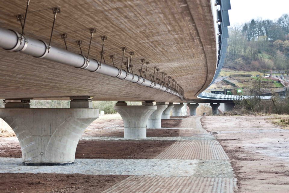 Unterseite einer Brücke mit mehreren Betonpfeilern, an der Metallrohre und Kabel entlanglaufen; darunter teils befestigter, teils unbefestigter Boden, im Hintergrund Bäume und Vegetation.