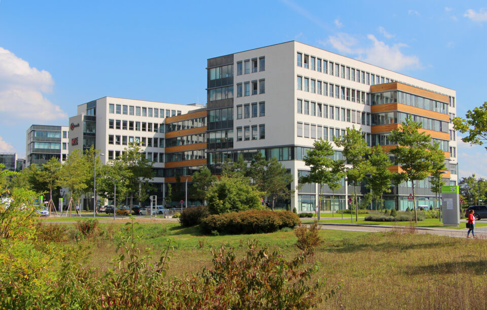 Modernes Bürogebäude mit mehreren Etagen, großen Fenstern und orangefarbenen Fassadenelementen, umgeben von Bäumen und Grünflächen unter leicht bewölktem Himmel; rechts geht eine Person entlang.