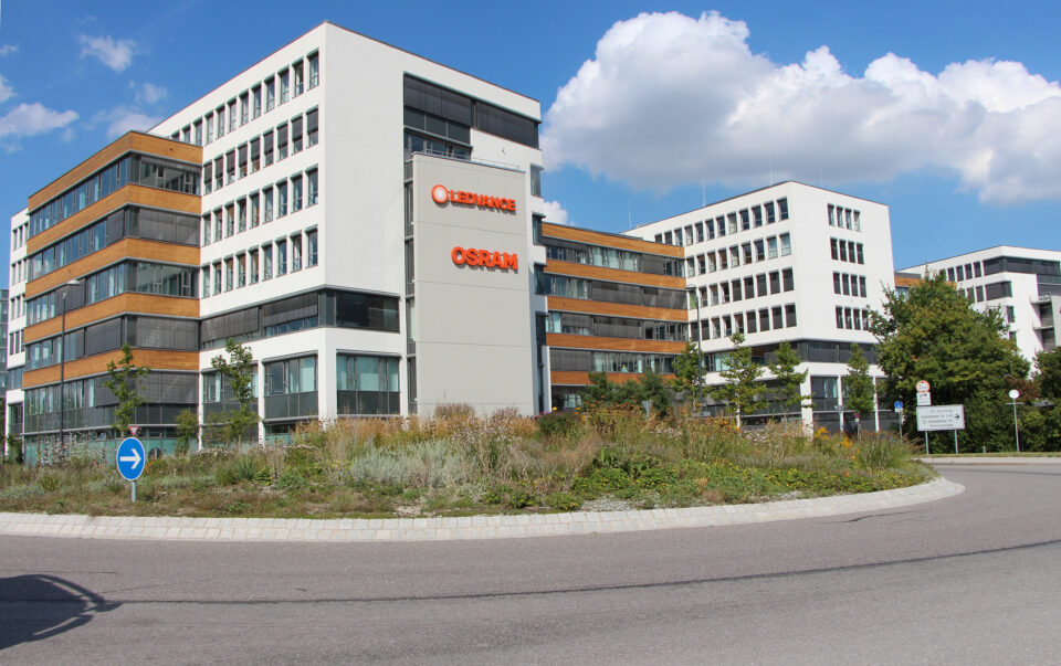 Moderner Bürokomplex mit weißen und holzverkleideten Fassaden, großen Fenstern und umliegender Begrünung; davor ein Kreisverkehr mit blauem Verkehrsschild