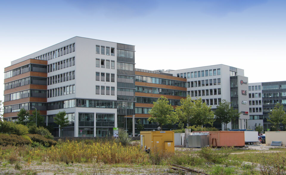 Moderner Bürokomplex mit mehreren verbundenen Gebäuden in Weiß- und Grautönen mit horizontalen Holzelementen, großen Fenstern, umgeben von Bäumen, Grünflächen und Baucontainern im Vordergrund.