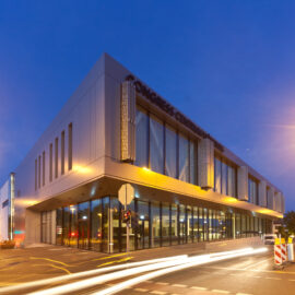 Modernes Gebäude mit der Aufschrift CONGRESS CENTRUM an der Fassade, abends oder nachts aufgenommen und beleuchtet; im Vordergrund Straßenlaternen und Lichtspuren vorbeifahrender Fahrzeuge in urbaner Umgebung.