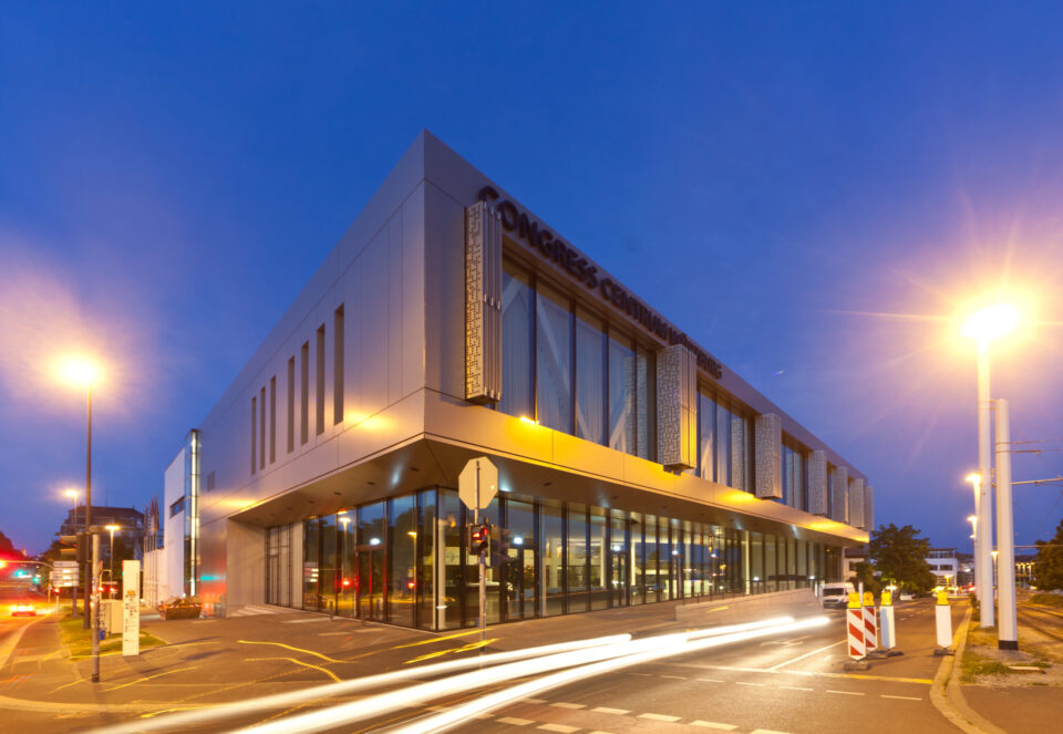 Modernes Gebäude mit der Aufschrift CONGRESS CENTRUM an der Fassade, abends oder nachts aufgenommen und beleuchtet; im Vordergrund Straßenlaternen und Lichtspuren vorbeifahrender Fahrzeuge in urbaner Umgebung.