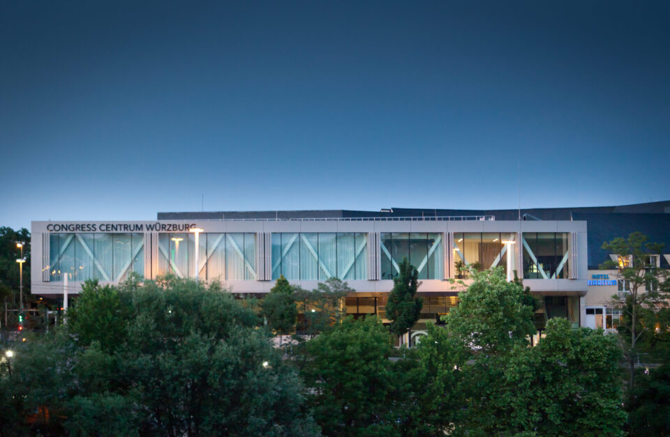 Congress Centrum Würzburg, modernes Gebäude mit großen Glasfenstern und elegantem Design, umgeben von Bäumen und beleuchtet von Straßenlaternen in den frühen Morgen- oder Abendstunden.