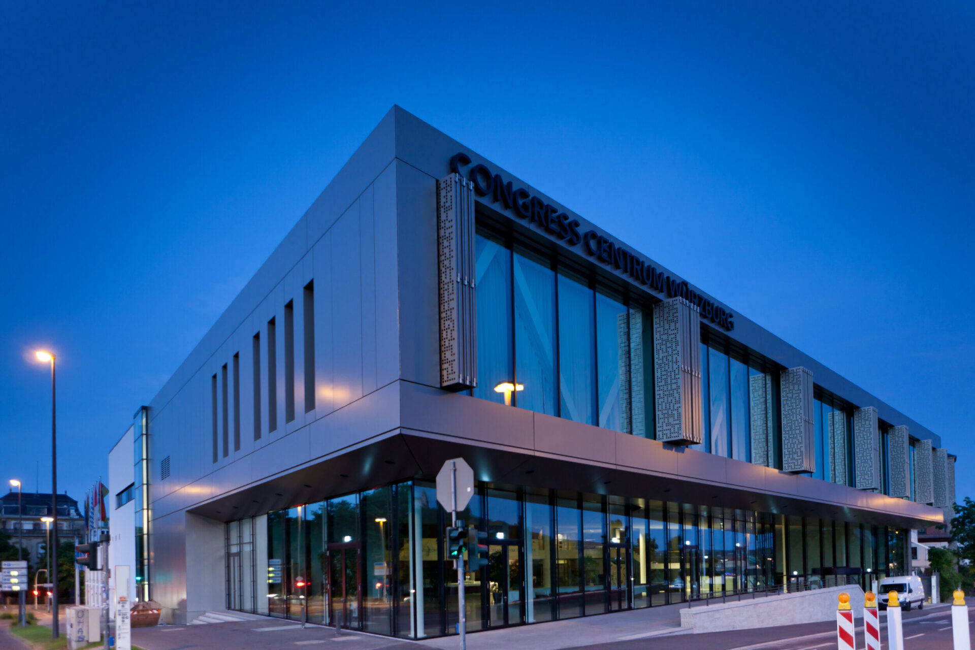 Modernes Gebäude mit der Aufschrift CONGRESS CENTRUM WÜRZBURG an der Fassade, mit großen Glasfenstern und zeitgemäßem Design; aufgenommen in der Dämmerung oder am frühen Abend, beleuchtet von Straßenlaternen.