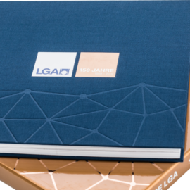 Das Bild zeigt ein Buch und darunter eine edle Buchhülle. Das Buch ist versehen mit einem geprägtem geometrischem Muster und einem Etikett mit der Aufschrift „LGA“ und „150 JAHRE“. Die Hülle darunter ist bronze mit weißer Schrift auf dem Buchrücken.