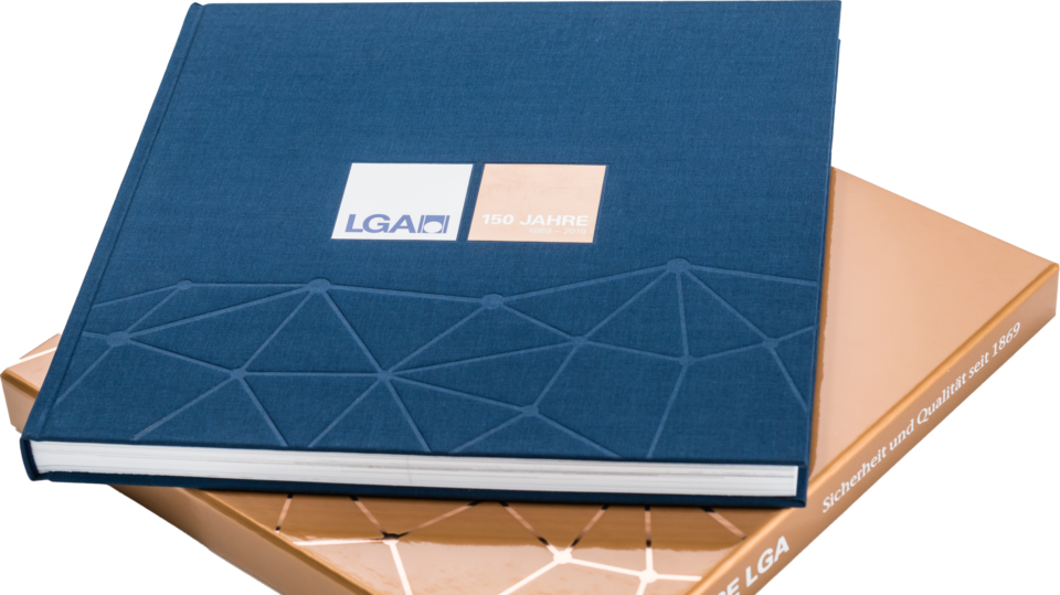 Das Bild zeigt ein Buch und darunter eine edle Buchhülle. Das Buch ist versehen mit einem geprägtem geometrischem Muster und einem Etikett mit der Aufschrift „LGA“ und „150 JAHRE“. Die Hülle darunter ist bronze mit weißer Schrift auf dem Buchrücken.