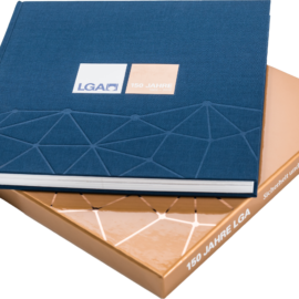 Das Bild zeigt ein Buch und darunter eine edle Buchhülle. Das Buch ist versehen mit einem geprägtem geometrischem Muster und einem Etikett mit der Aufschrift „LGA“ und „150 JAHRE“. Die Hülle darunter ist bronze mit weißer Schrift auf dem Buchrücken.