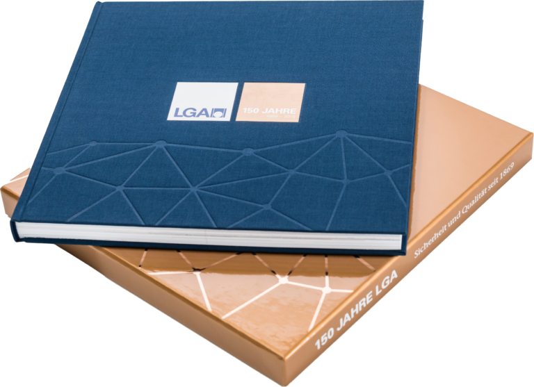 Das Bild zeigt ein Buch und darunter eine edle Buchhülle. Das Buch ist versehen mit einem geprägtem geometrischem Muster und einem Etikett mit der Aufschrift „LGA“ und „150 JAHRE“. Die Hülle darunter ist bronze mit weißer Schrift auf dem Buchrücken.