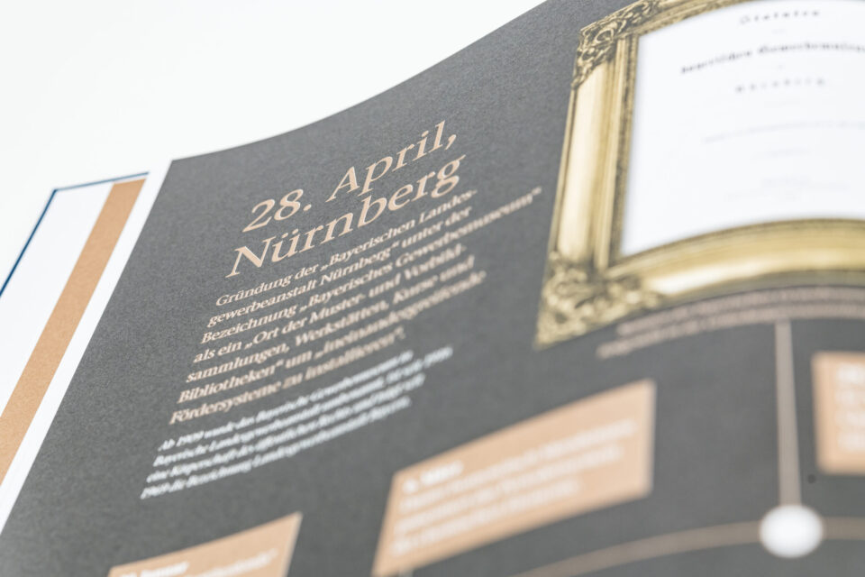 Das Bild zeigt ein aufgeklapptes Buch mit einer Doppelseite. Auf der linken Seite befindet sich ein Text zur Gründung der Bayerischen Landesgewerbeanstalt in Nürnberg, auf der rechten Seite ein gerahmtes historisches Dokument.