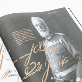 Das Bild zeigt ein aufgeschlagenes Buch mit einer Schwarz-Weiß-Fotografie eines älteren Mannes in Militäruniform. Nebenan befindet sich ein erklärender Text zur Rolle von König Ludwig III. im Bezug auf die LGA Landesgewerbeanstalt Bayern s sowie eine große handschriftliche Unterschrift.