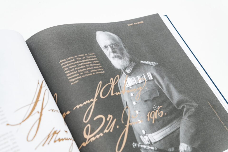 Das Bild zeigt ein aufgeschlagenes Buch mit einer Schwarz-Weiß-Fotografie eines älteren Mannes in Militäruniform. Nebenan befindet sich ein erklärender Text zur Rolle von König Ludwig III. im Bezug auf die LGA Landesgewerbeanstalt Bayern s sowie eine große handschriftliche Unterschrift.