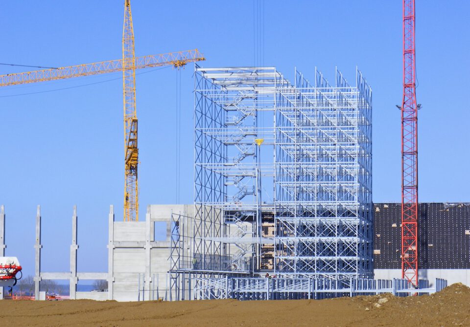 Eine Baustelle mit einem teilweise errichteten Stahlbau, unterstützt von einem gelben und einem roten Kran; sichtbar sind Stahlträger, Betonelemente und Baumaterialien auf unbefestigtem Boden unter klarem, blauem Himmel.