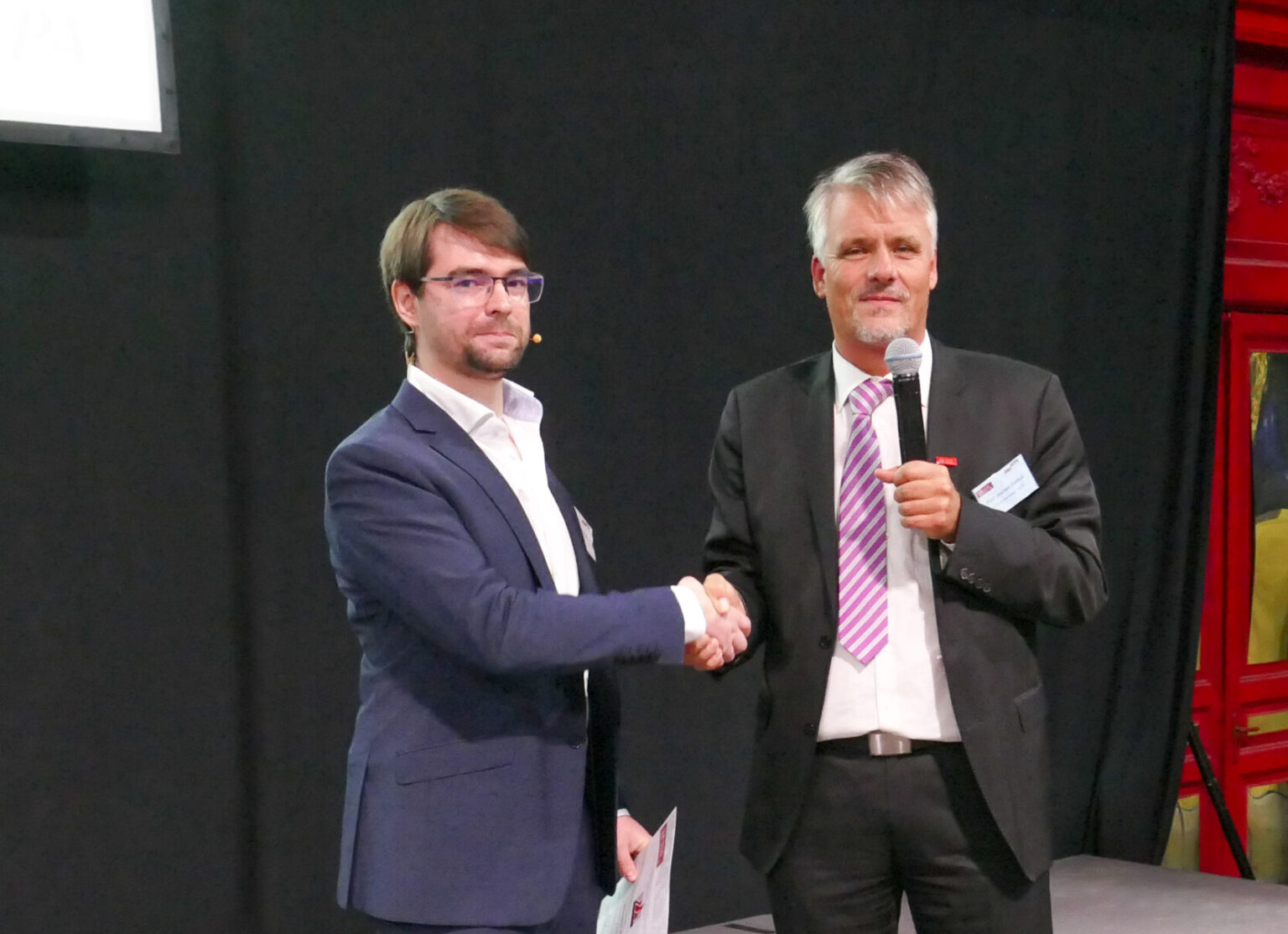 Zwei Personen in formeller Kleidung geben sich die Hand; eine hält ein Mikrofon, die andere ein Blatt Papier, vor dunklem Hintergrund.