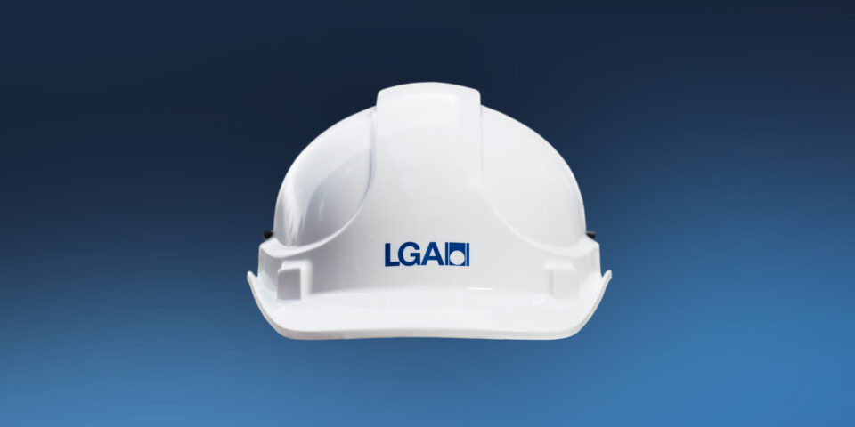Ein weißer Schutzhelm mit dem Logo „LGA“ ist zentral im Bild zu sehen. Der Hintergrund verläuft von dunklem Blau oben zu hellem Blau unten.