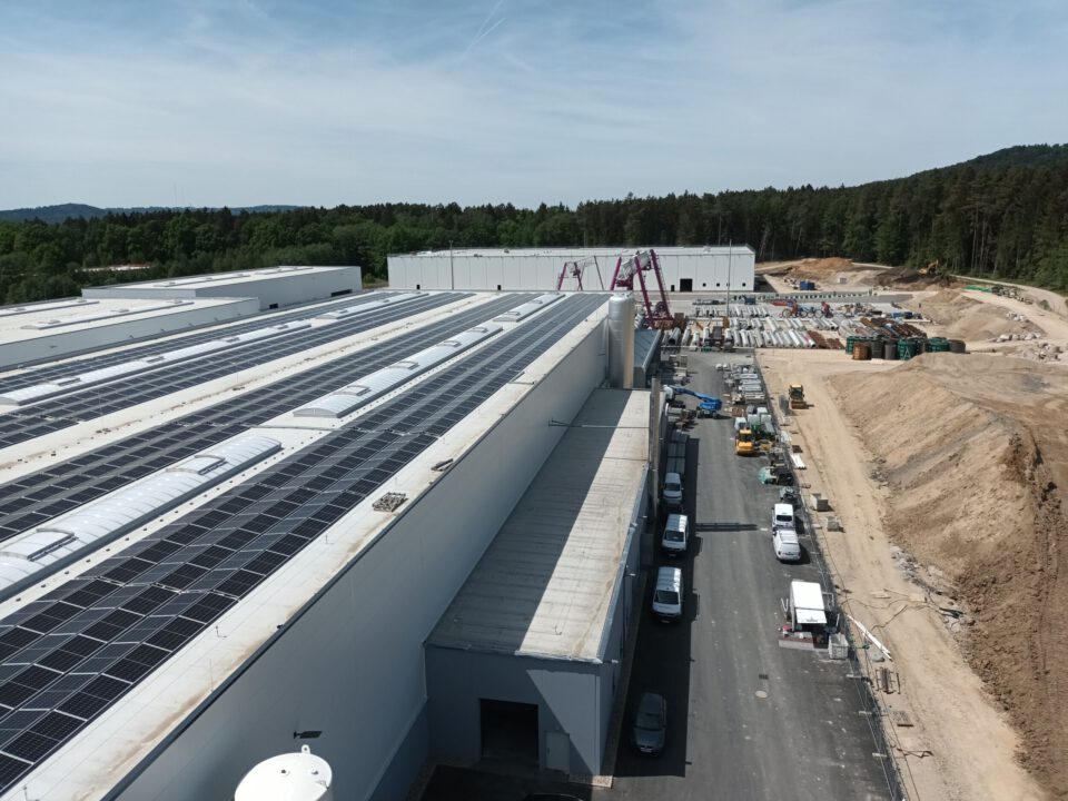Luftaufnahme eines großen Industriekomplexes mit mehreren Gebäuden, deren Dächer mit Solarpanels bedeckt sind, angrenzender Straße mit geparkten Fahrzeugen sowie Baumaterialien und Baumaschinen im Hintergrund inmitten einer grünen Umgebung.