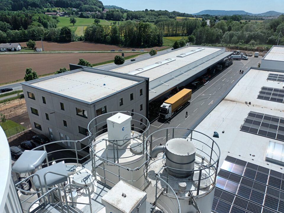 Industrieareal mit mehreren Gebäuden, darunter ein großes Lager mit Laderampe und geparkten Lkw, ein mehrstöckiges Gebäude mit Fenstern sowie Industrieanlagen mit Tanks und Rohrleitungen im Vordergrund; im Hintergrund grüne Felder und Hügel unter klarem Himmel.