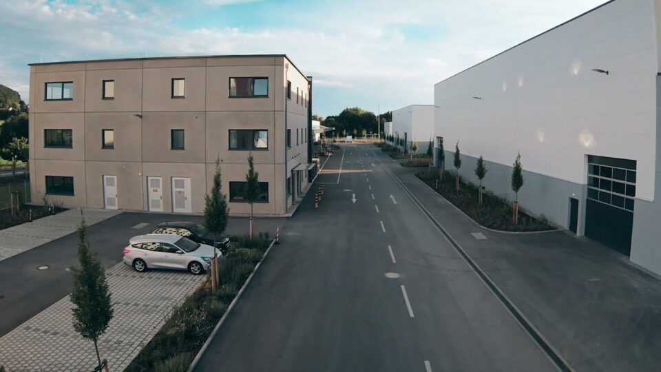 Straße in einem Industrie- oder Gewerbegebiet mit einem dreistöckigen Gebäude und geparktem Auto auf der linken Seite sowie großen weißen Hallen und kleinen Bäumen auf der rechten Seite unter teils bewölktem Himmel.
