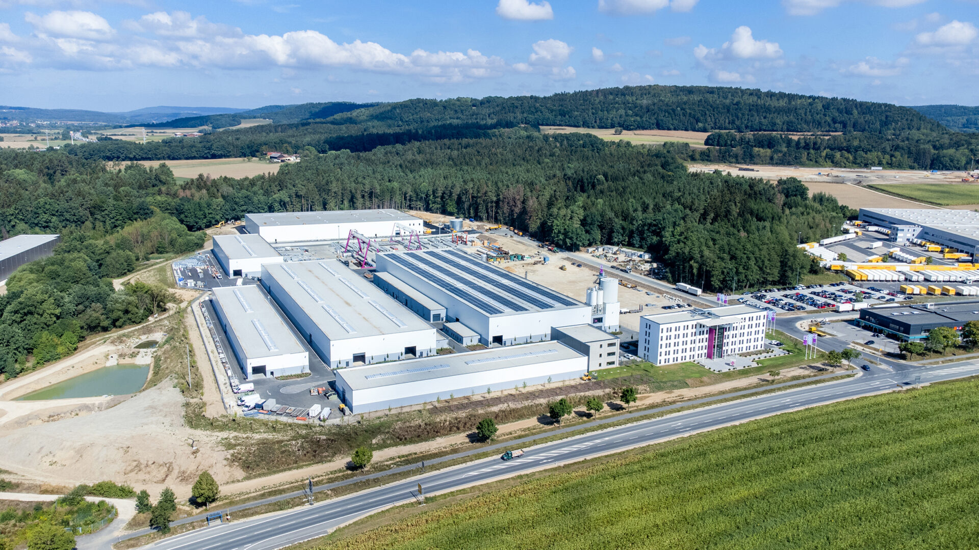 Luftaufnahme eines großen Industriekomplexes mit mehreren weißen Hallen, einem Bürogebäude und Baustellenbereichen, umgeben von Feldern, Wald und einer Straße mit Fahrzeugen im Vordergrund.