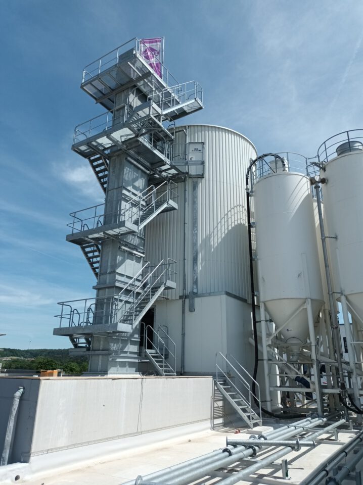 Industrieanlage mit großen weißen Zylindertanks und einer Metalltreppe, die zu einer Plattform mit Fahne führt, vermutlich zur Wartung oder Überwachung der Tanks, unter leicht bewölktem Himmel.