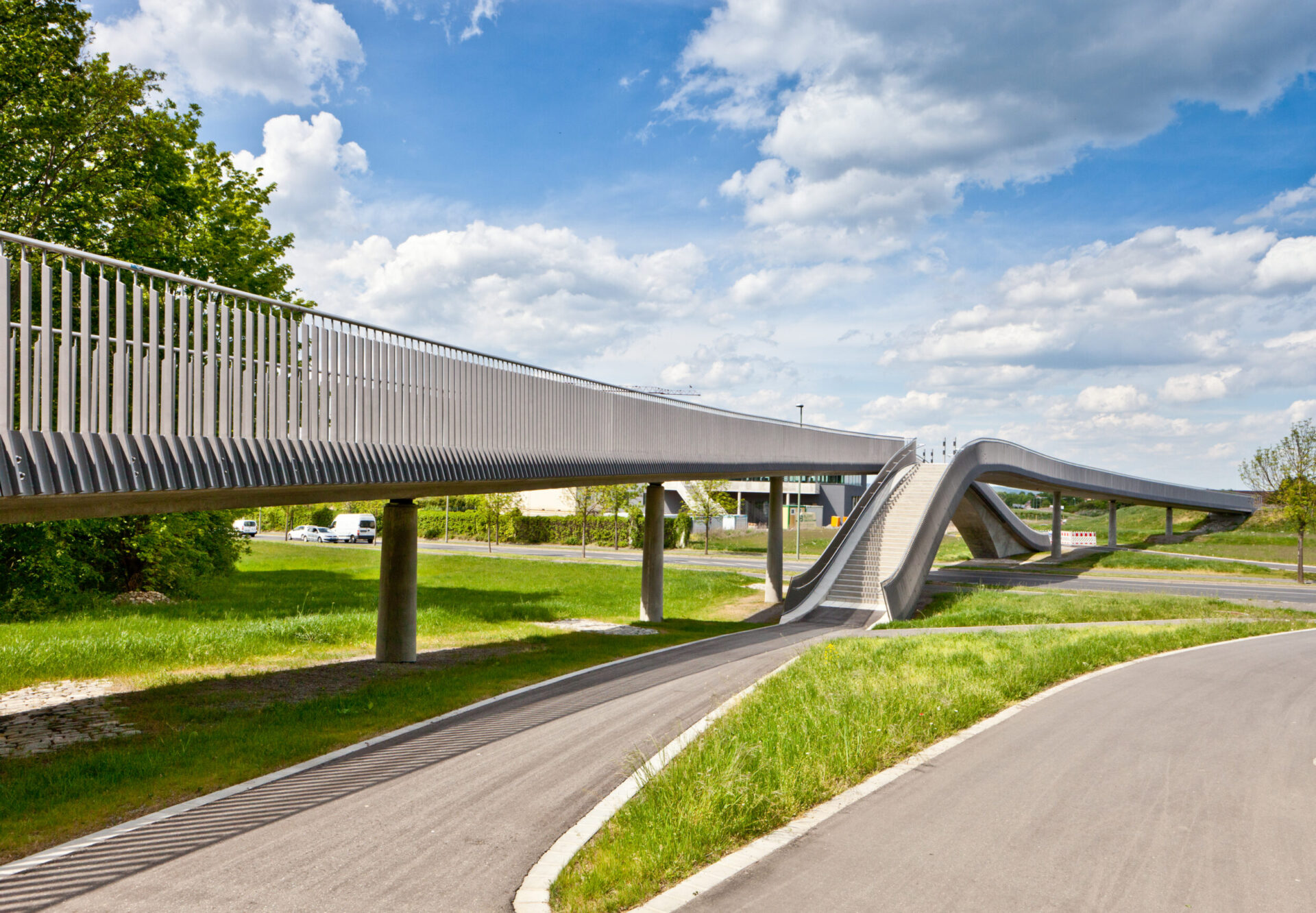 Eine moderne Fußgängerbrücke mit wellenförmigem Design verläuft auf Betonpfeilern über eine Straße, umgeben von grüner Wiese, Bäumen und einem blauen Himmel mit vereinzelten Wolken.