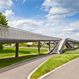 Eine moderne Fußgängerbrücke mit wellenförmigem Design verläuft auf Betonpfeilern über eine Straße, umgeben von grüner Wiese, Bäumen und einem blauen Himmel mit vereinzelten Wolken.