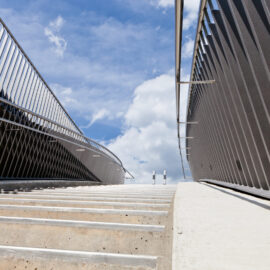 Eine Betontreppe mit Metallgeländern auf beiden Seiten führt nach oben in Richtung eines hellblauen Himmels mit vereinzelten Wolken, aufgenommen aus der Perspektive am Fuß der Treppe.