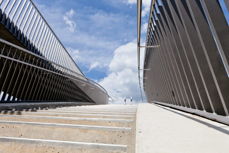 Eine Betontreppe mit Metallgeländern auf beiden Seiten führt nach oben in Richtung eines hellblauen Himmels mit vereinzelten Wolken, aufgenommen aus der Perspektive am Fuß der Treppe.