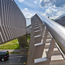 Eine Fußgängerbrücke mit Metallgeländern und Treppen führt über eine Straße, auf der ein schwarzes Auto fährt, unter einem teilweise bewölkten Himmel.