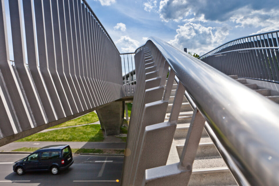 Eine Fußgängerbrücke mit Metallgeländern und Treppen führt über eine Straße, auf der ein schwarzes Auto fährt, unter einem teilweise bewölkten Himmel.