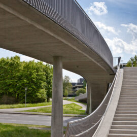 Eine erhöhte Fußgängerbrücke mit Treppenaufgang verläuft auf Betonpfeilern über eine Straße, im Hintergrund sind Bäume, Grünflächen und ein teilweise bewölkter Himmel mit blauen Abschnitten zu sehen.