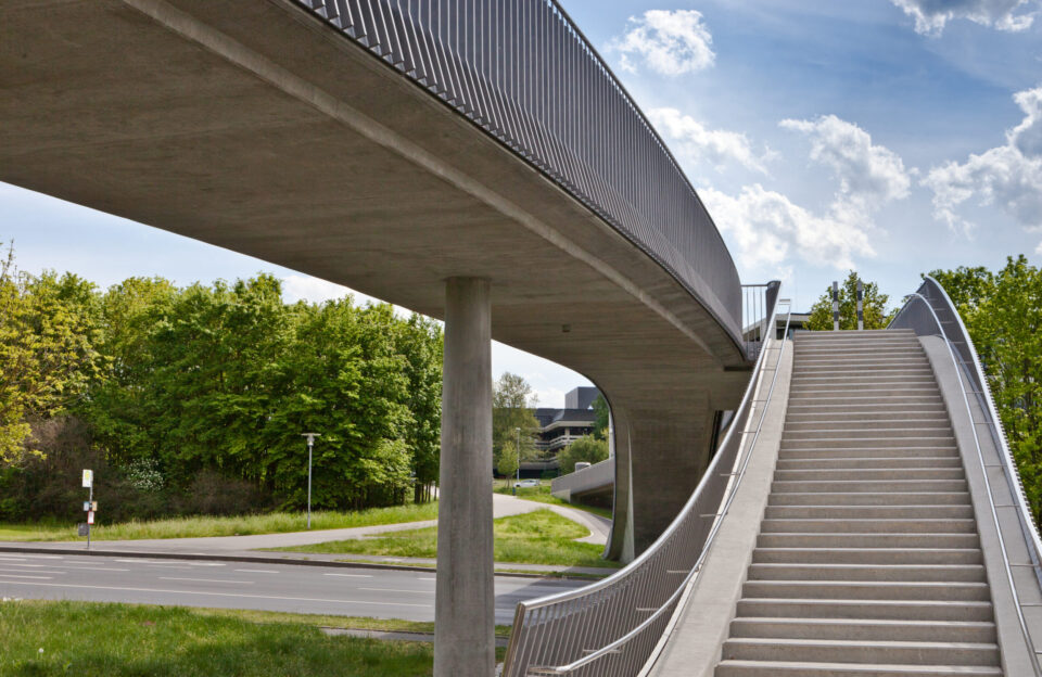 Eine erhöhte Fußgängerbrücke mit Treppenaufgang verläuft auf Betonpfeilern über eine Straße, im Hintergrund sind Bäume, Grünflächen und ein teilweise bewölkter Himmel mit blauen Abschnitten zu sehen.
