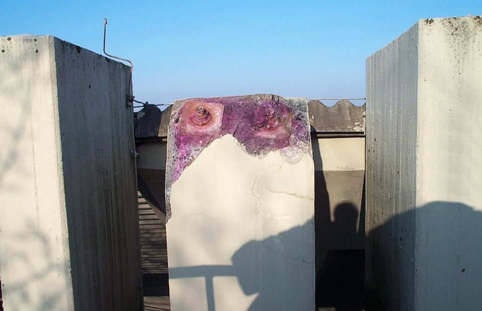 Drei große Betonsäulen ragen vor blauem Himmel empor, wobei die mittlere Säule Verfärbungen und rostige Schrauben am oberen Ende aufweist; Sonnenlicht wirft deutliche Schatten auf die Strukturen.