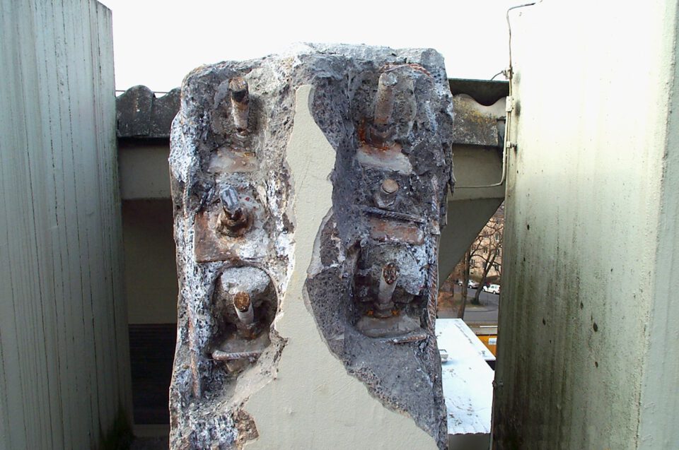 Nahaufnahme einer beschädigten Betonstruktur mit freiliegenden, verrosteten Bewehrungsstäben; der Beton ist stellenweise abgeplatzt und gibt den Blick auf die korrodierten Metallteile frei.