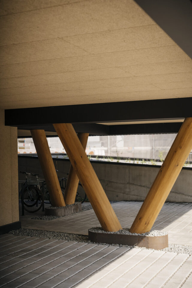 Gebäudebereich mit V-förmig angeordneten Holzträgern, die auf Betonsockeln stehen, umgeben von Kies und Betonplattenboden; im Hintergrund ist teilweise ein Fahrrad sichtbar, die Decke hat eine strukturierte Oberfläche und natürliches Licht fällt von einer Seite ein.
