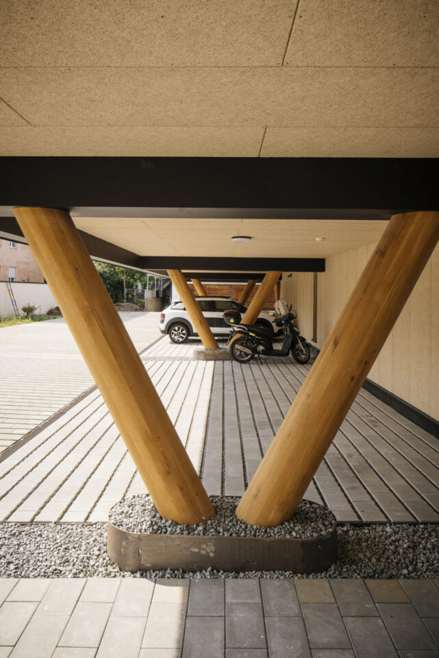 Überdachter Parkplatz mit V-förmig angeordneten Holzstützen, unter denen ein weißes Auto und ein Motorrad stehen; der Boden ist mit rechteckigen Platten gepflastert, um die Stützen herum befinden sich Kiesflächen, die Decke besteht aus hellem Material.