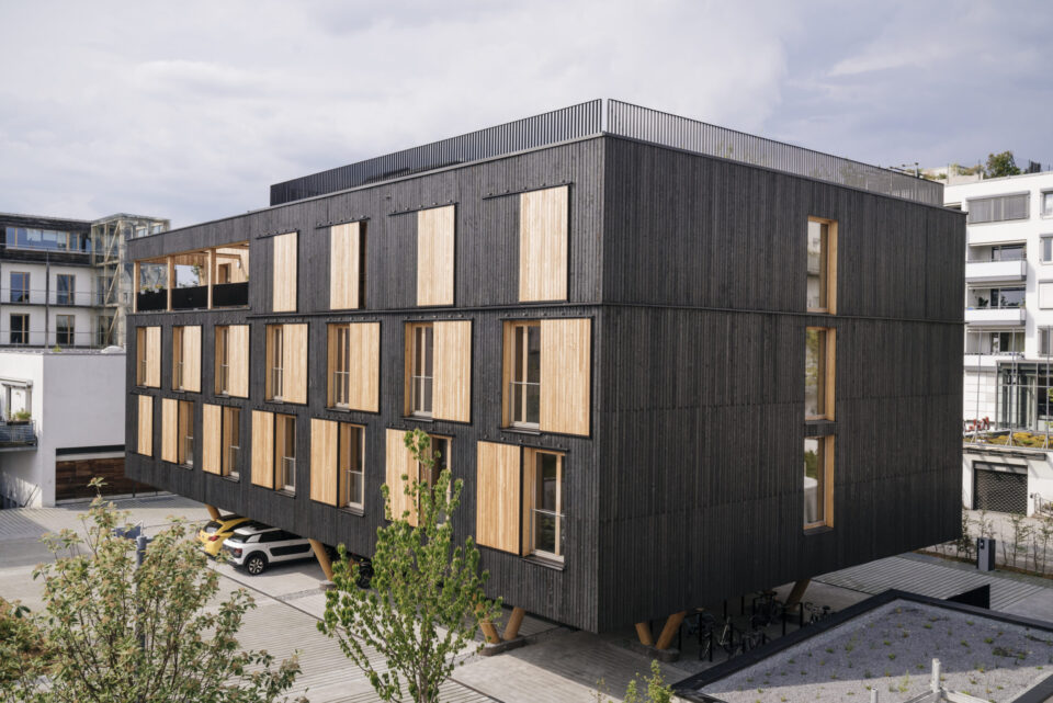 Modernes, rechteckiges Gebäude mit dunkler Fassade und Holzfensterläden, drei Etagen mit mehreren Fenstern pro Seite, davor ein kleiner Parkplatz mit einem unter dem Gebäude geparkten Auto; im Hintergrund Bäume und weitere Gebäude.