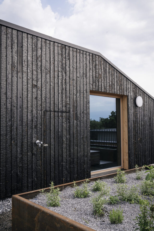 Modernes Gebäude mit dunkler, strukturierter Holzfassade und rechteckiger Tür mit Holzrahmen auf der rechten Seite; davor ein Hochbeet mit Kies und kleinen grünen Pflanzen, unter teils bewölktem Himmel.