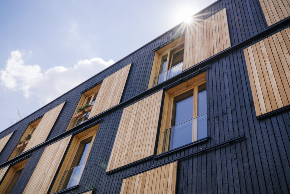 Außenansicht eines modernen Gebäudes mit einer Kombination aus dunklen und hellen Holzpaneelen, großen Fenstern und teils verglasten Geländern, beleuchtet von Sonnenlicht aus der rechten oberen Ecke.