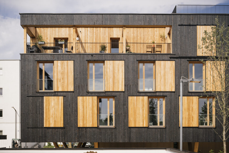 Moderne Gebäudefassade mit einer Kombination aus hellen Holzpaneelen und dunkler vertikaler Verkleidung, drei Etagen, wobei das oberste Stockwerk einen offenen Balkon mit Pflanzen und Gartenmöbeln aufweist; die mittleren und unteren Etagen haben große Fenster mit Holzrahmen.