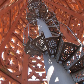 Das Bild zeigt eine Wendeltreppe aus Metall in einem kunstvoll gestalteten Bauwerk mit filigranen Mustern an den Wänden, durch die Licht einfällt, wobei sich die Treppe um eine zentrale Säule nach oben windet.