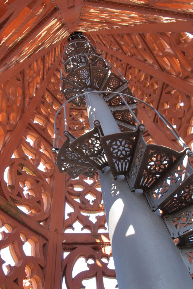 Das Bild zeigt eine Wendeltreppe aus Metall in einem kunstvoll gestalteten Bauwerk mit filigranen Mustern an den Wänden, durch die Licht einfällt, wobei sich die Treppe um eine zentrale Säule nach oben windet.