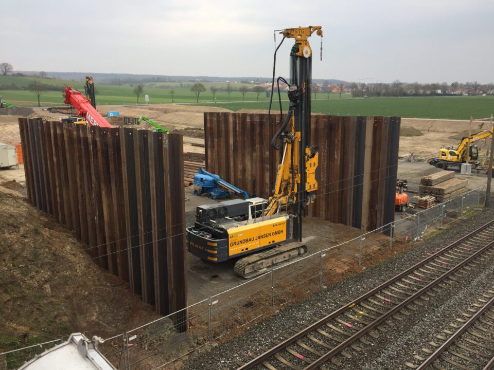 Baustelle mit großen Stahlspundwänden, die ein rechteckiges Baufeld umschließen; darin befinden sich schwere Baumaschinen, darunter ein gelber Bohrwagen sowie Kräne und Bagger, angrenzend an Bahngleise mit grünen Feldern und Gebäuden im Hintergrund.