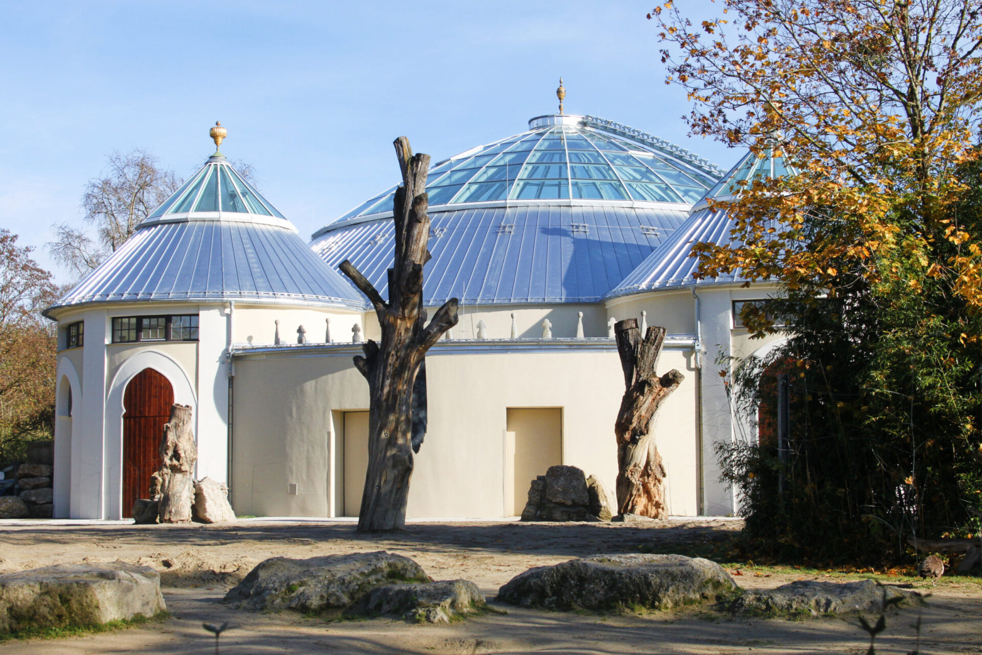 Gebäude mit markanter Architektur und drei gläsernen Kuppeldächern – einer großen zentralen und zwei kleineren seitlichen; im Vordergrund Baumstümpfe, Felsen und herbstlich gefärbte Bäume unter klarem Himmel.