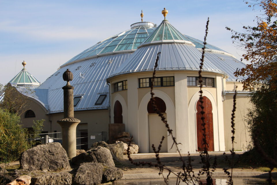 Großes, kuppelförmiges Gebäude mit metallischem Dach und goldenen Verzierungen, mehreren hölzernen Rundbogentüren und -fenstern, umgeben von Felsen, Pflanzen und einer Steinsäule im Vordergrund bei klarem Himmel.